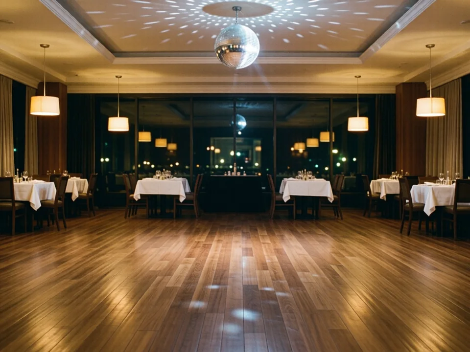 Symbolbild: Leere polierte Tanzfläche in einem Restaurant bei Nacht, ringsum eingedeckte Tische mit weißen Decken, warmes Licht von Pendelleuchten und eine Spiegelkugel, die kleine Lichtpunkte im Raum verteilt.